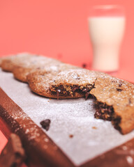 Freshly made Chocolate Chip Cookies on a wooden serving board. Cookies with a glass of fresh milk on the side along with chocolate chips and pieces of chocolate around on a pink background.Freshly mad
