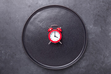 Red alarm clock and empty black ceramic plate on dark stone background. Intermittent fasting concept