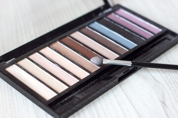 An eyes shadow palette and a matte brush in nude beige shades that have the appearance of being used on a wooden light background