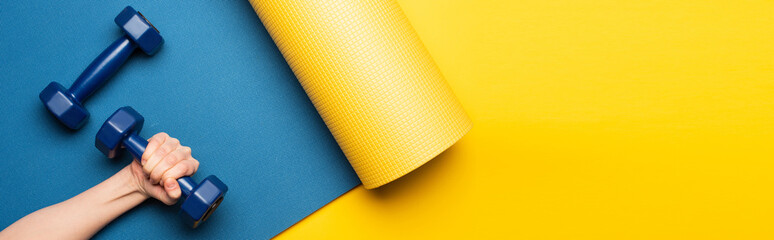cropped view of woman holding dumbbell on blue fitness mat on yellow background, panoramic shot