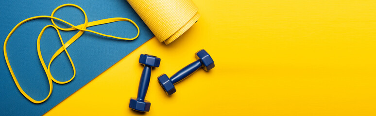 top view of blue fitness mat with dumbbells and resistance band on yellow background, panoramic shot