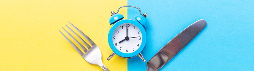 Blue alarm clock, fork, knife on colored paper background. Intermittent fasting concept. Horizontal banner