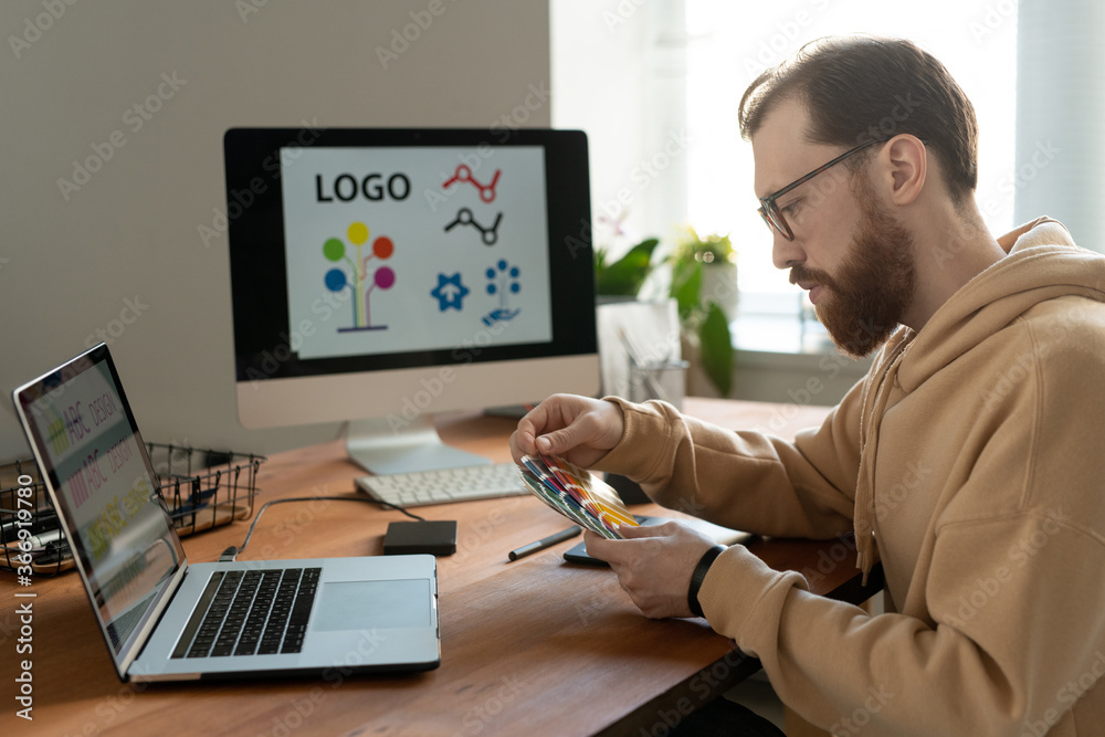 Wall mural serious bearded designer in eyeglasses sitting at desk with computers and viewing color swatch while