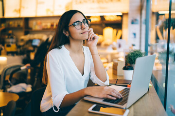 Pensive caucasian female in casual wear and spectacles thinking about creative ideas for blog publication, dreamy businesswoman thinking about vacations and recreation during remote job in cafe
