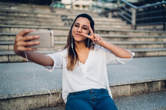 Positive beautiful caucasian woman in eyewear and earphones making selfie on smartphone camera, happy female blogger posing for picture on mobile phone share multimedia content in social networks