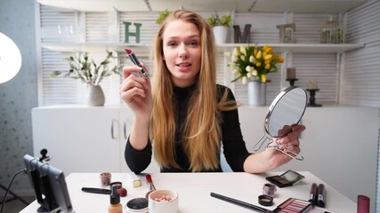 Vlogger female applies lipstick on lips. Beauty blogger woman filming daily makeup routine tutorial at camera on tripod. Influencer blonde girl live streaming cosmetics product review in home studio