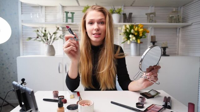 Vlogger female applies lipstick on lips. Beauty blogger woman filming daily makeup routine tutorial at camera on tripod. Influencer blonde girl live streaming cosmetics product review in home studio