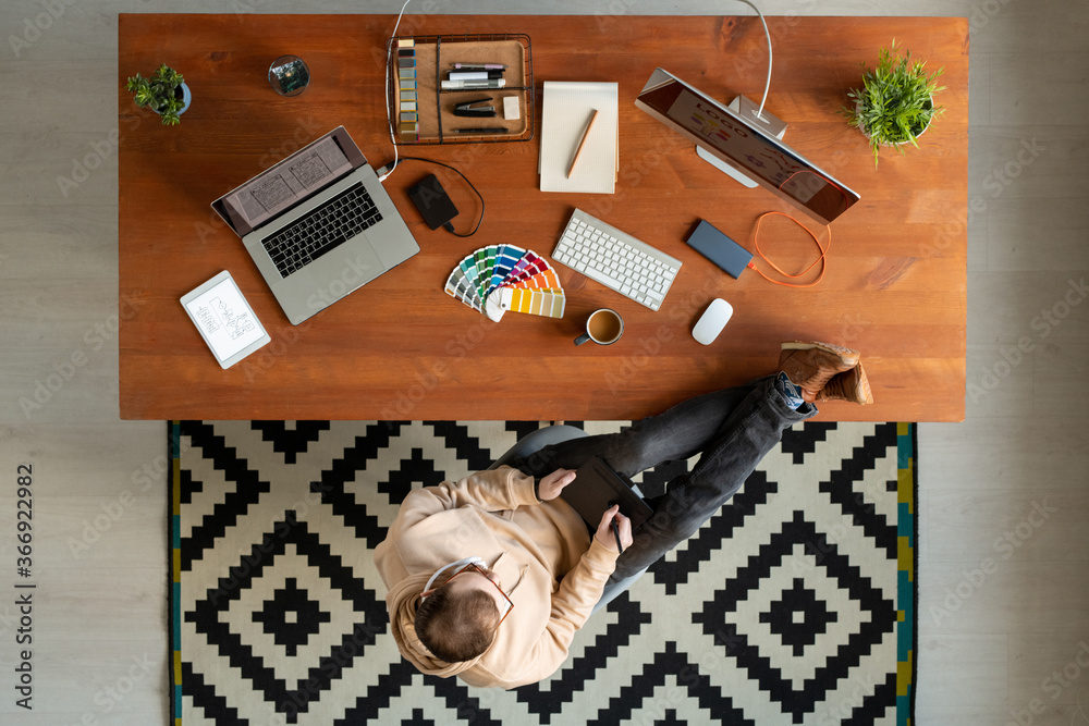 Wall mural directly above view of young man keeping feet on table and using drawing pad while working on graphi
