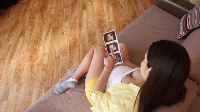 A pregnant woman is looking at ultrasound scans while sitting on the couch. Anticipating the birth of a child. Family values. Stroke your tummy.