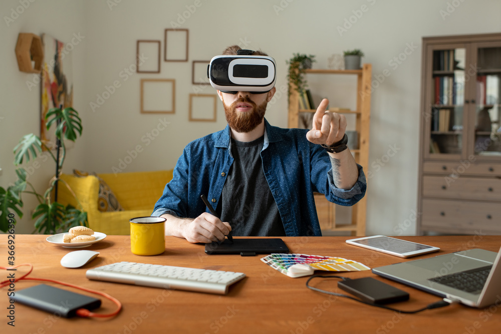 Wall mural serious bearded designer sitting at table and drawing on digitizer while using virtual reality simul