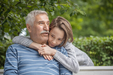 portrait of grandson and grandfather, teenager boy hugs his grandfather in the park on a bench, summer vacation with grandfather