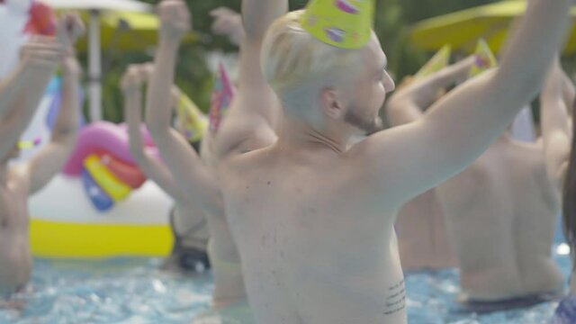 Close-up of cheerful young tattooed man with blond dyed hair dancing and spinning in water at resort. Bearded Caucasian animator having fun with people celebrating holiday in pool.