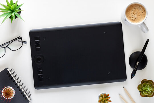 A Graphics Tablet In The Center Of A White Desk. Office Utensils All Around. Coffee, Notebook, Succulents.