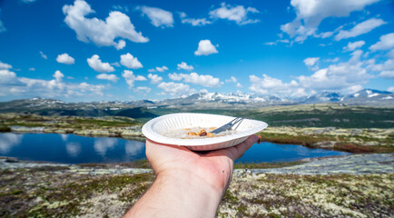 Hand holding a bowl of oatmeal outdoors in nature with sunny and warm weather. Mountain scenery and blue sky.