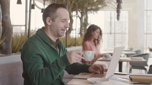 Pan shot of Caucasian middle-aged man and his female colleague sitting at tables in modern coworking space, drinking tea and using laptops