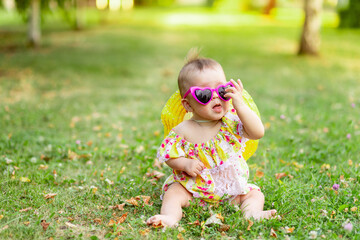 a small baby girl of 7 months is sitting on the green grass in a yellow dress, hat and bright glasses and showing her tongue, walking in the fresh air. Space for text