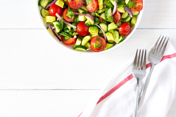 Salad of fresh ripe tomato, onion, avocado, cucumber and parsley and olive oil, salad in a white plate with a fork and towel on a white wooden background