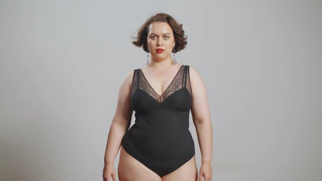 Beautiful brunette plus-size woman in black sexy swimsuit posing on grey background