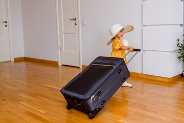 A little happy girl in oversize hat and yellow dress is carrying a big suitcase in room with white walls and wood floor