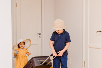 Happy girl and boy, brother and sister  are carrying a big suitcase in room with white walls and wood floor
