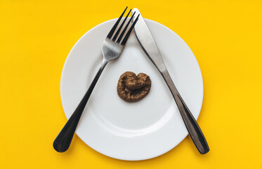 Plastic excrement of lies on a white plate with fork and knife on yellow tablecloths. The concept of an inept cook, fake news, a crappy restaurant, bad food, a taste of shit. Bad smelling joke.