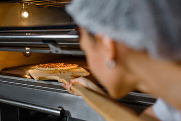 Young caucasian woman baker is holding a wood peel with fresh pizza and put it in an oven at a...
