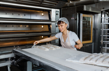 Young caucasian woman baker is preparing bread dough and cut it with knife for bread baker process in electric oven at baking manufacture factory