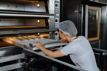 Young caucasian woman baker is putting the bread dough and in electric oven at baking manufacture factory.