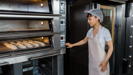 Young caucasian woman baker is looking to the bread baker proces and turn on the electic oven at...
