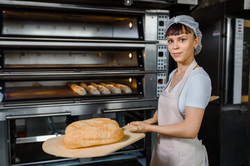 Young caucasian woman baker is holding a wood peel with fresh bread near an oven at baking manufacture factory.