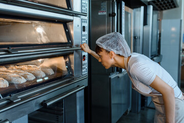 Young caucasian woman baker is looking at the bread baker process in an electric oven at a baking manufacturing factory.