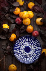 yellow ripe plums and empty ceramic plate among leaves on rustic old wooden table background