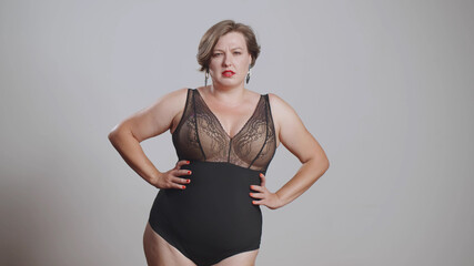 Plump young woman in black body stockings standing on grey background with hands on hips