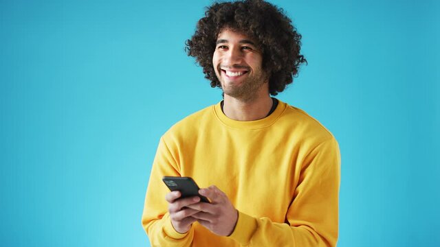 Studio portrait of confident smiling young man looking at mobile phone against blue background - shot in slow motion