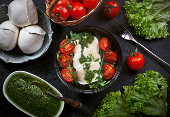 fresh mozzarella salad with lettuce and arugula leaves, pesto sauce, fresh cherry tomatoes and cedar nuts, on dark slate table background