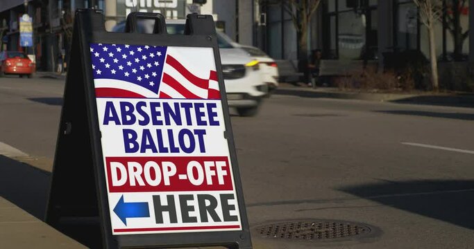 A sign on the sidewalk of a large city shows where to drop off absentee ballots.  	