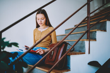 Thoughtful brunette hipster girl sitting on stairs in campus with trendy leather backpack checking email on smartphone,student reading interesting book and chatting with friend in networks in collage