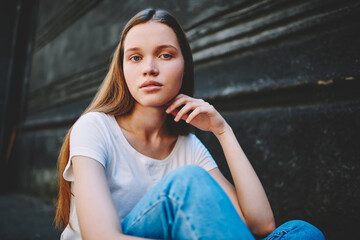 Portrait of beautiful brunette young woman in casual outfit sitting on wall background looking at camera,attractive hipster girl with natural makeup resting on city street enjoying free time