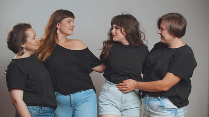 Cheerful plus size women in black t-shirts and jeans embracing and smiling at camera