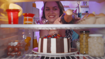 View from refrigerator of beautiful stout woman taking champagne from fridge at home party