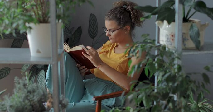 Woman reading a book in room of plants. Young florist in eyeglasses sitting on armchair and reading a book.