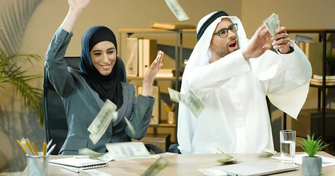 Arabian young businesswoman and businessman in traditional Middle Eastern clothes counting money and tossing banknotes. Muslim rich wife and husband throwing and playing with dollars bills in office.