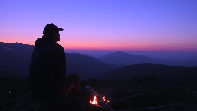A man sits by a fire in the mountains and looks at the night sky. Campfire in the tent camp at night. Hiking in the mountains.