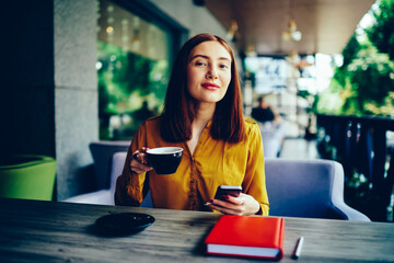 Half length portrait of positive young hipster girl with piercing in nose having recreation time with gadget and tasty coffee.Young pretty woman booking online on website using smartphone and 4G