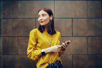 Positive redhead woman dressed in stylish clothing looking away while listening to favorite radio station via cellular and earphones.Happy female student relaxing with music during summer walk