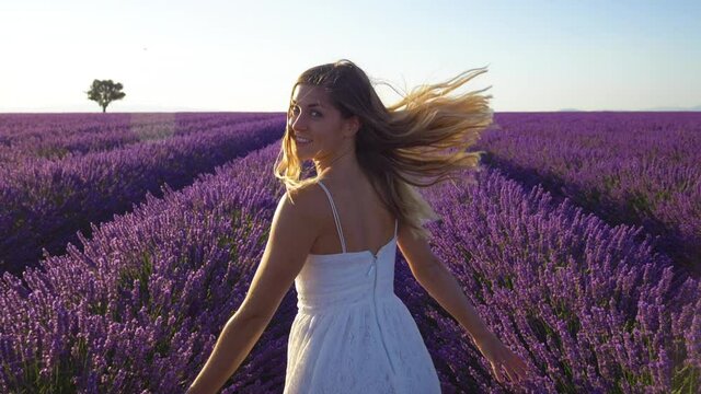 Slow motion - Happy young woman in a white dress running through lavender field and looking back into the camera