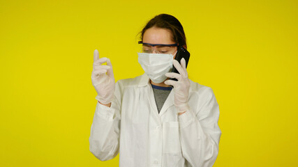 Young doctor in medical mask, goggles, latex gloves talking to patient on phone. Girl in white coat on yellow background holds smartphone near ear. Coronavirus, flu