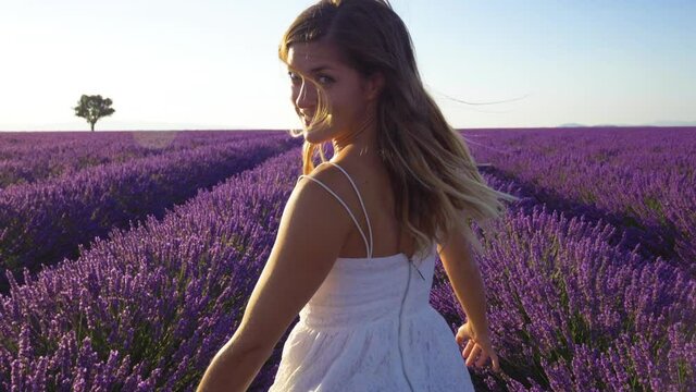 Slow motion - Happy young woman in a white dress running through lavender field and looking back into the camera