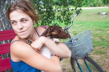 A girl in a blue sundress sits on a bench by a birches and holds a beautiful little brown and white dog in her arms.Portrait of a girl with a dog.Bicycle with a basket.Pet love, dog day, village scene
