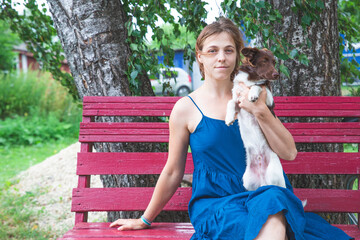 A girl in a blue sundress sits on a bench by the birches and holds a beautiful little brown and white dog in her arms. Portrait of a girl with a dog. Love for pets, dog day, rural scene